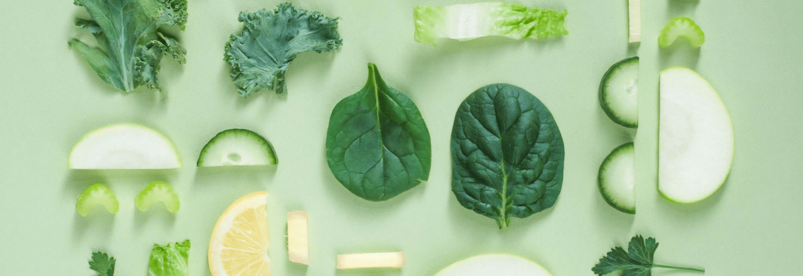 Groene bladgroenten en plakjes groenten op een groene achtergrond, waaronder boerenkool, spinazie, selderij, komkommer, en citroen.